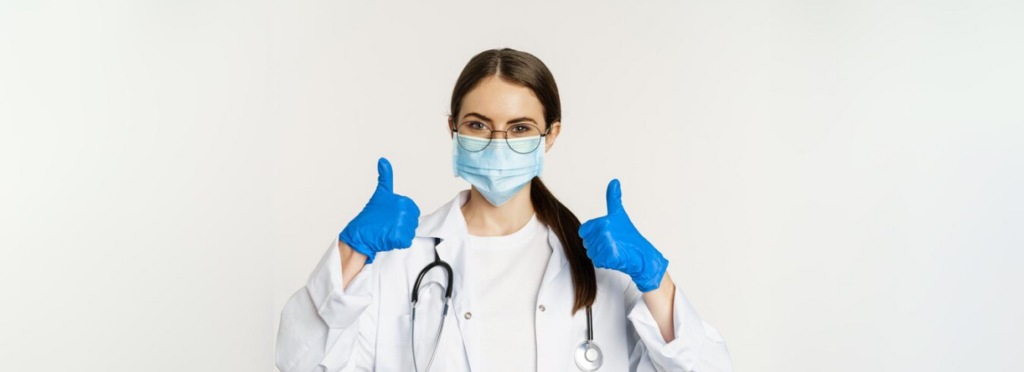 woman in blue gloves and mask showing thumbs up in front of white wall