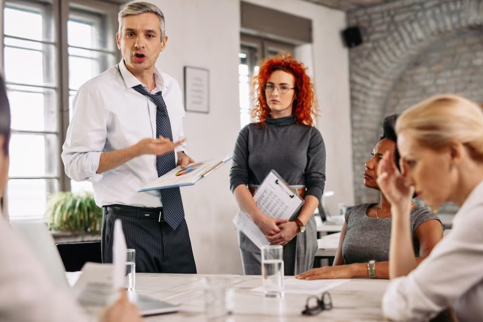 a man with papers in his hand and a woman are standing over their colleagues who are sitting