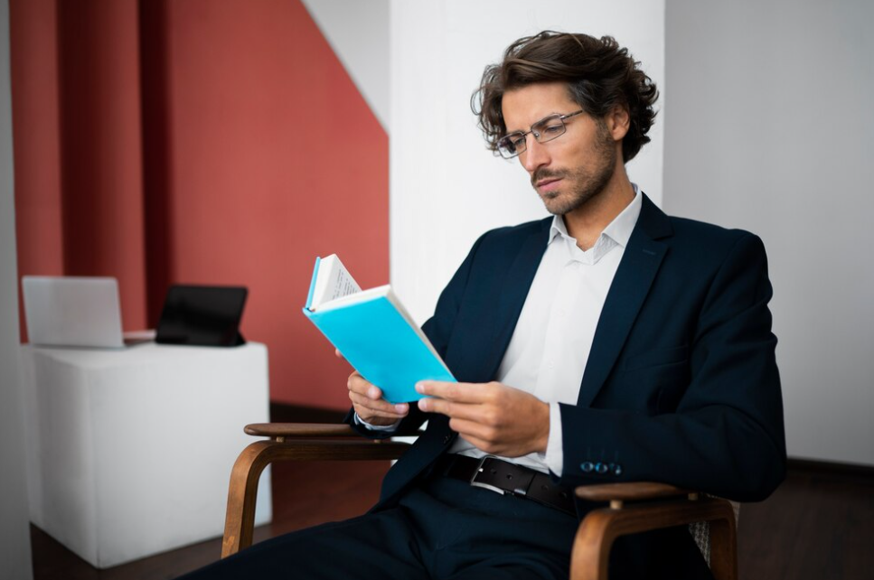 man in a classic suit and glasses sitting in a chair and reading the blue book